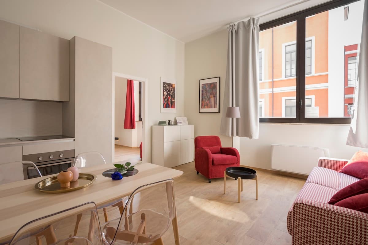 Monoambiente con cocina integrada, mesa con sillas transparentes, sillón rojo y ventana a un edificio naranja