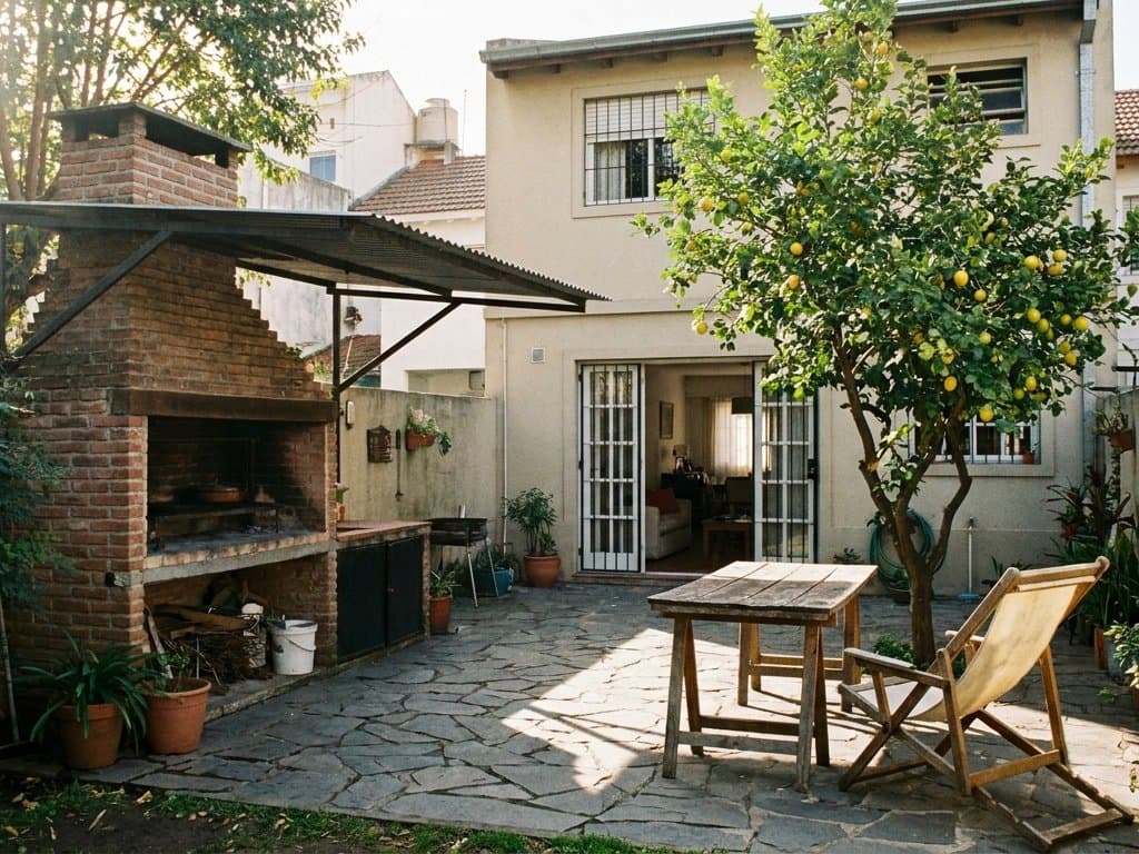 Patio de una casa con parrilla de ladrillo, mesa de madera, reposera y un limonero cargado de frutas